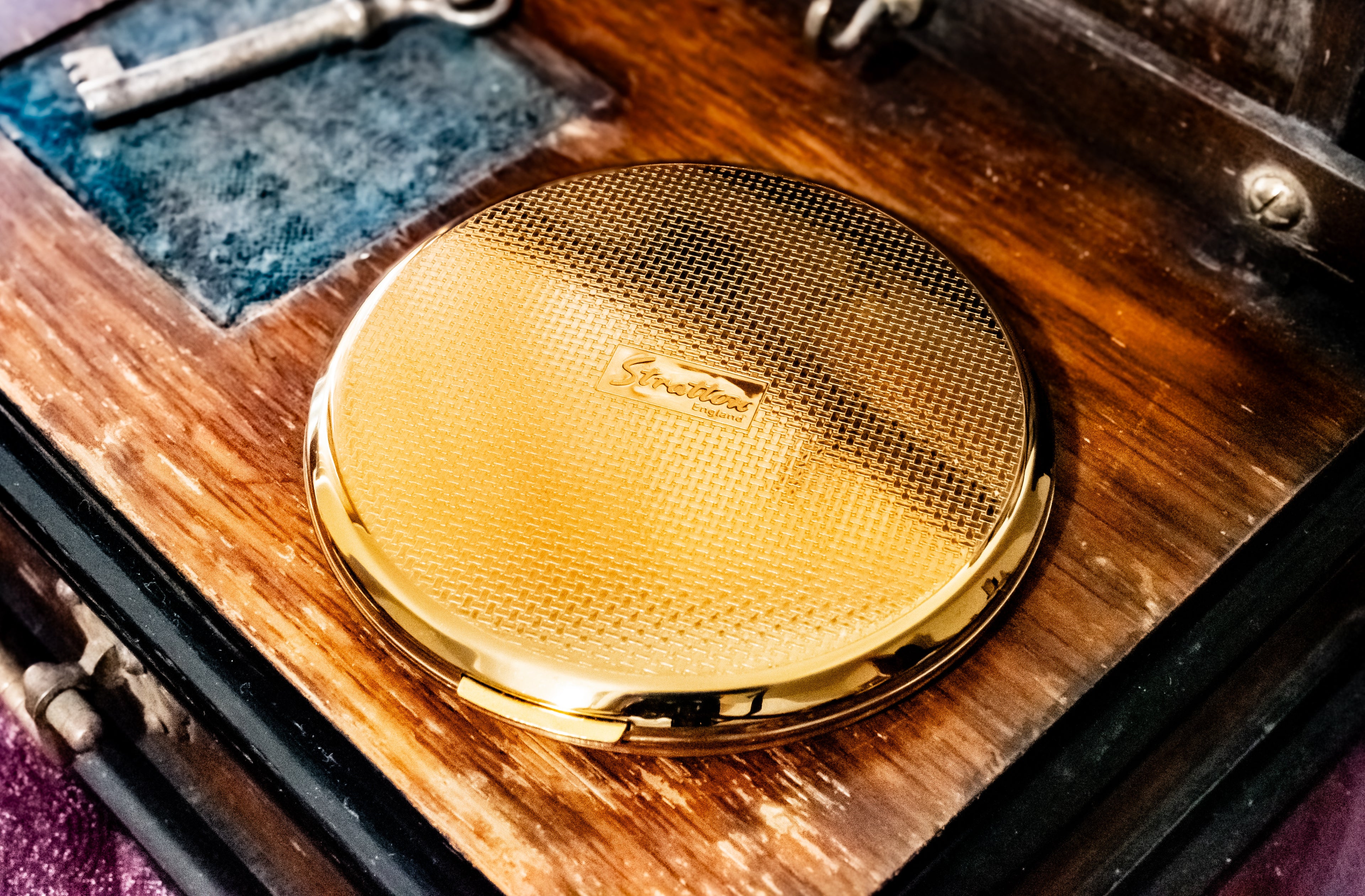 A round gold compact mirror with a textured honeycomb pattern lid and a small horse logo in the center, resting on a vintage wooden box with a blue velvet insert and an antique key.
