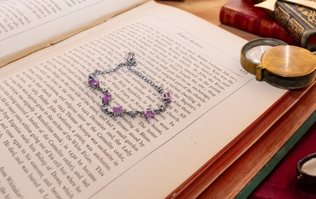 A silver-toned chain bracelet with purple gemstone accents rests on the pages of an open vintage book, surrounded by old books and a brass pocket mirror on a wooden surface.