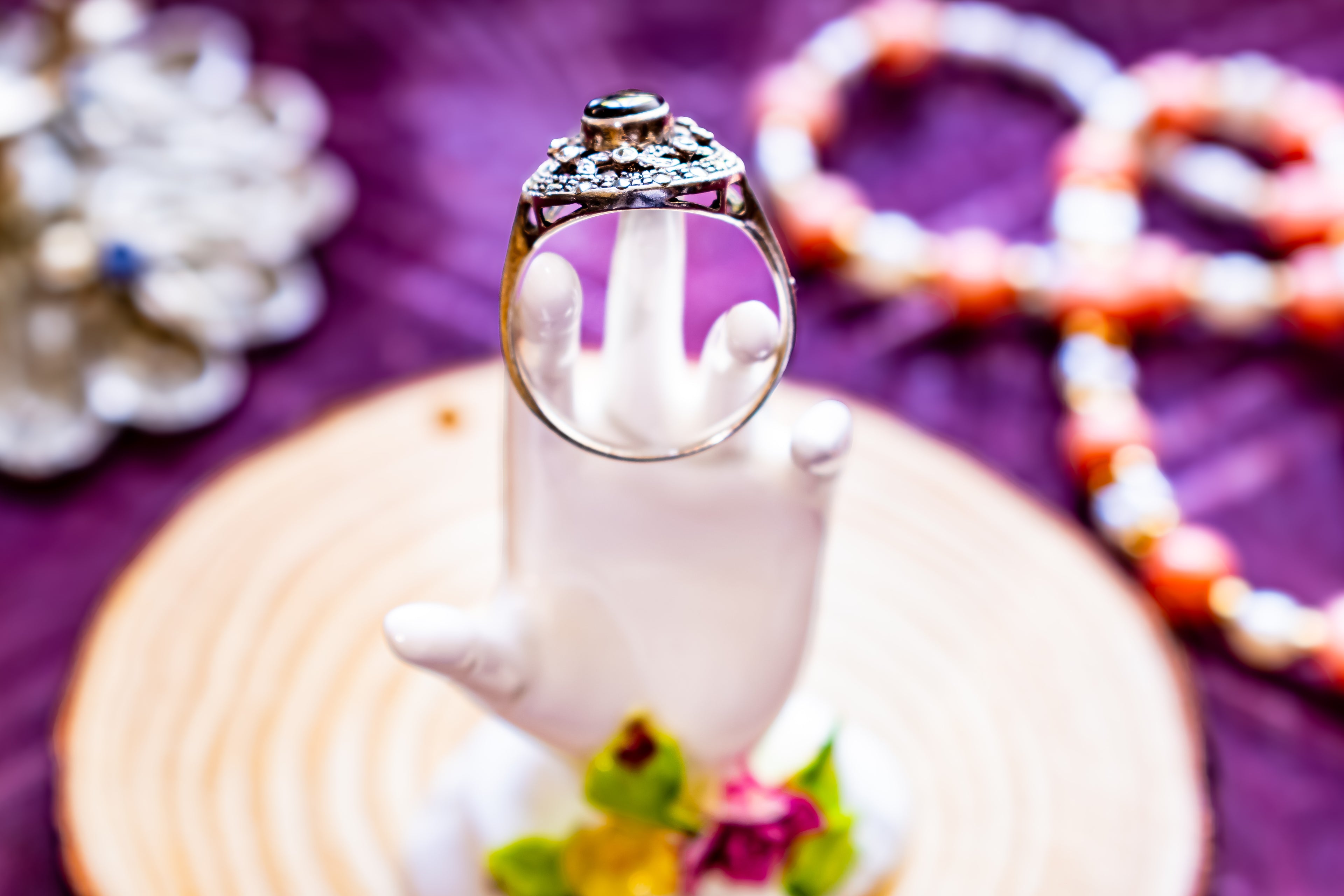A silver ring with a large central dark gemstone and intricate filigree detailing, displayed on a white hand-shaped stand. The stand is placed on a circular wooden slice over a purple fabric background, with blurred beads in the background.