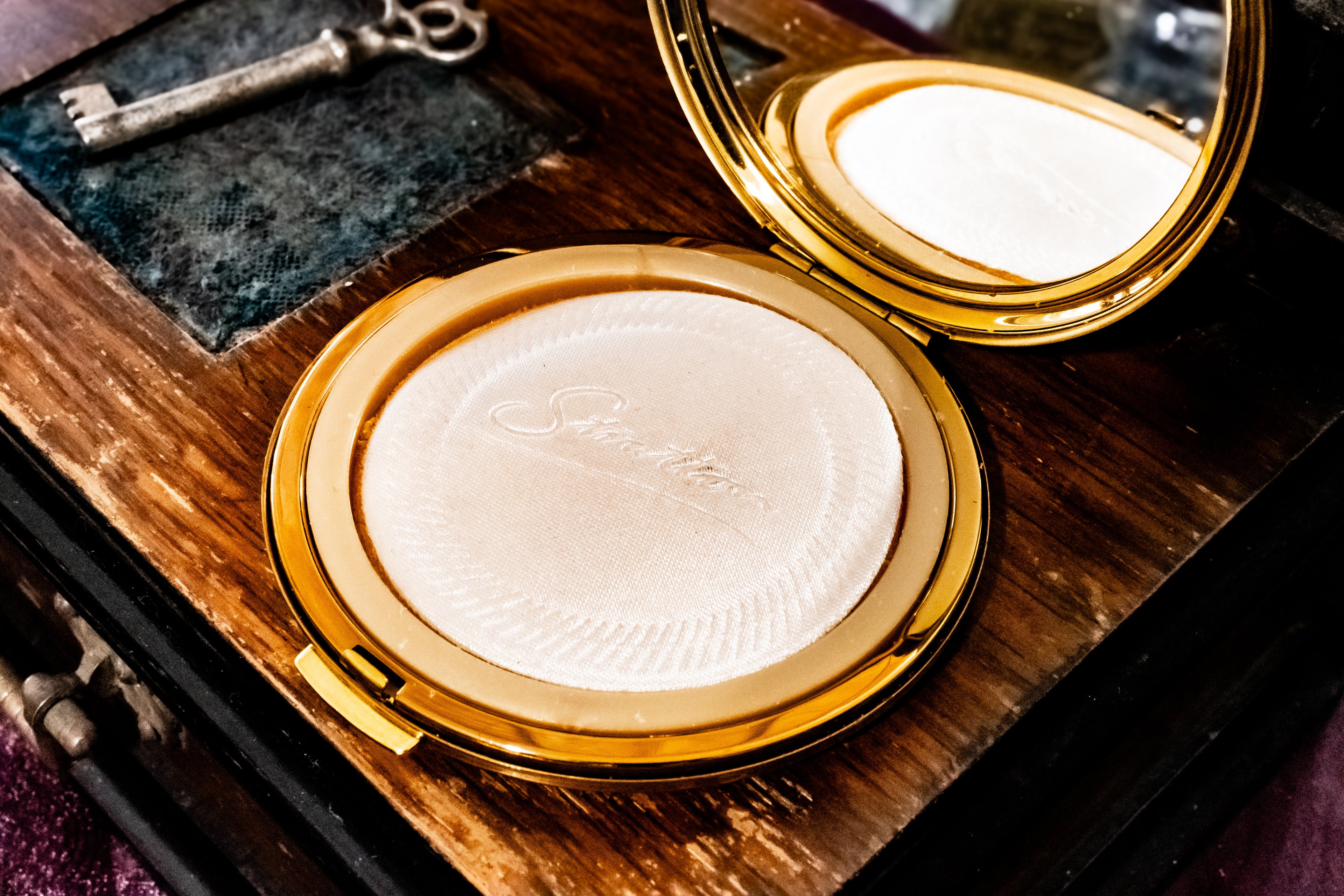 An open round gold compact containing a pressed powder with an embossed script logo, resting on a dark wooden surface next to a vintage-style key and a mirror.