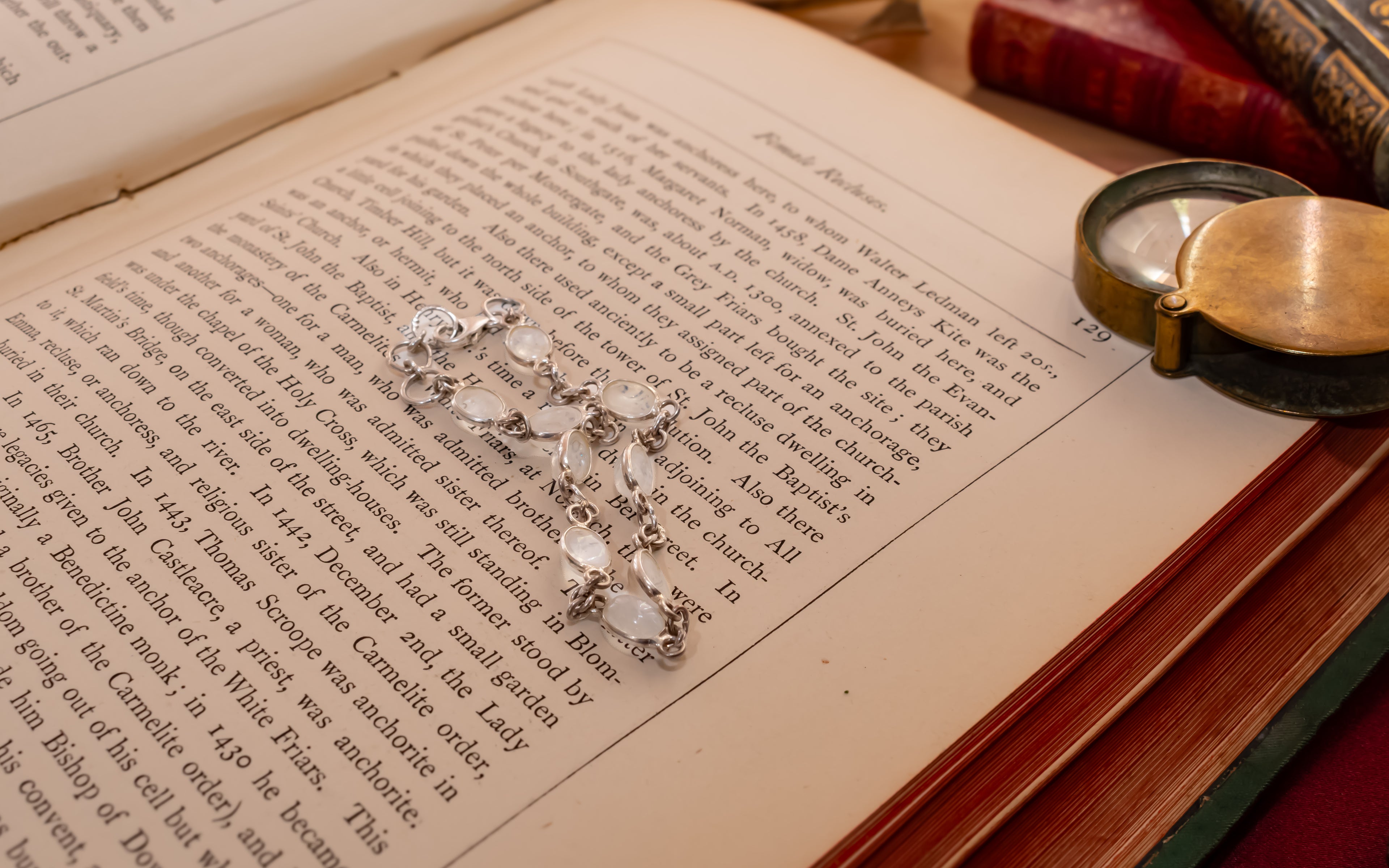 A silver-toned multi-stone pendant necklace with clear faceted crystals rests on the open pages of a vintage book, surrounded by old leather-bound books and a brass pocket mirror.