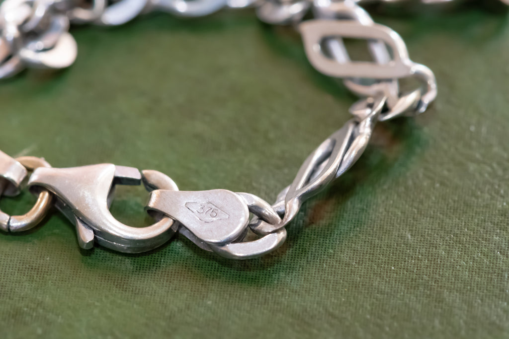 A close-up of a silver-toned metal chain bracelet with a lobster clasp and twisted links, resting on a textured olive green surface.