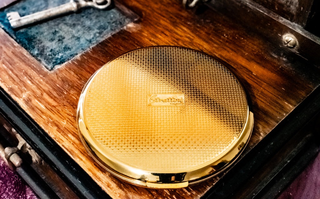 A round gold compact mirror with a textured honeycomb lid and a small central logo, resting on a dark wooden surface with an antique key in the background.