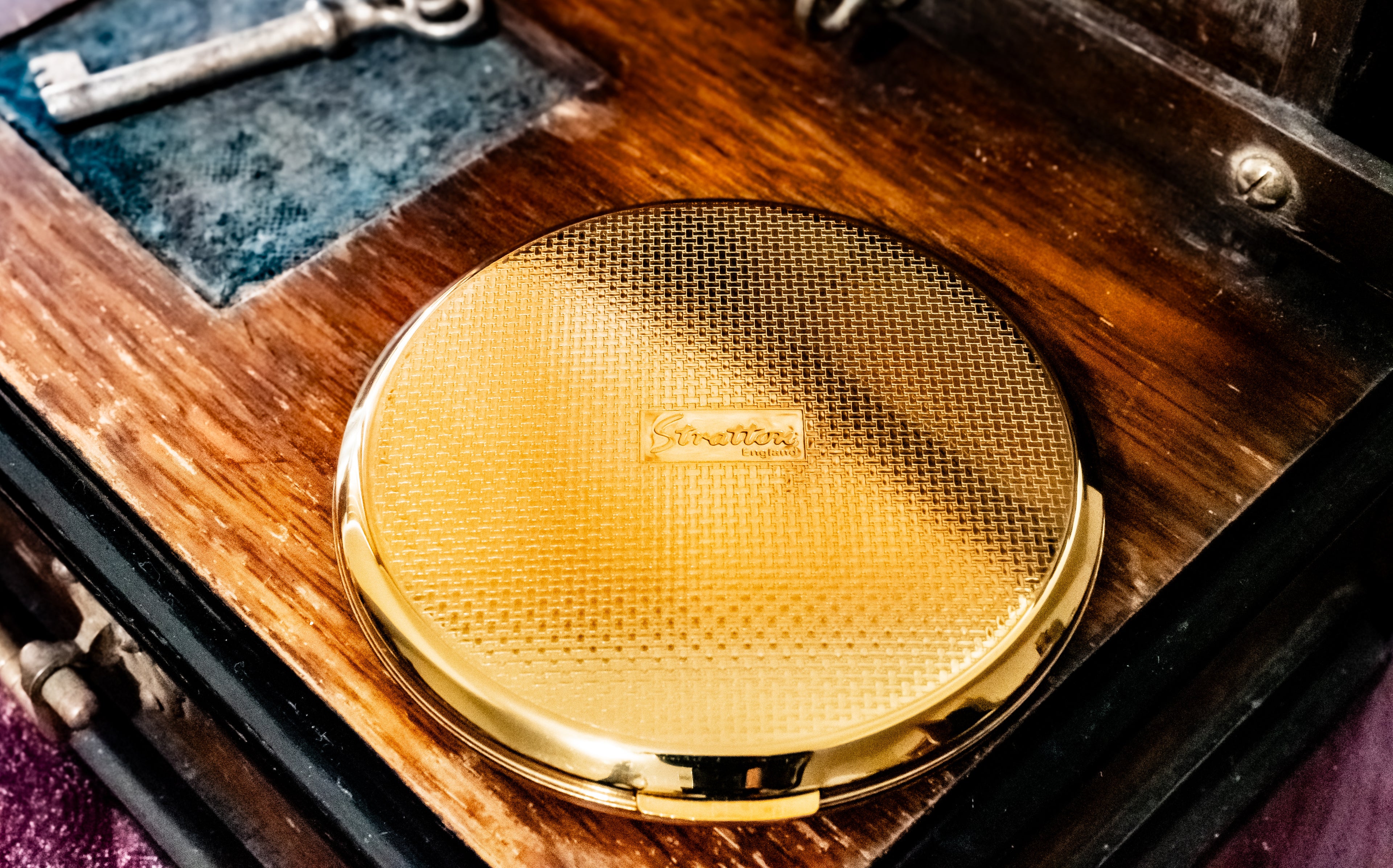 A round gold compact mirror with a textured honeycomb lid and a small central logo, resting on a dark wooden surface with an antique key in the background.