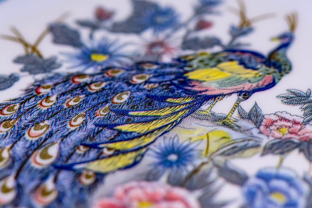 A close-up of a colorful embroidered peacock with blue, yellow, green, and pink details, surrounded by floral motifs on a white background.