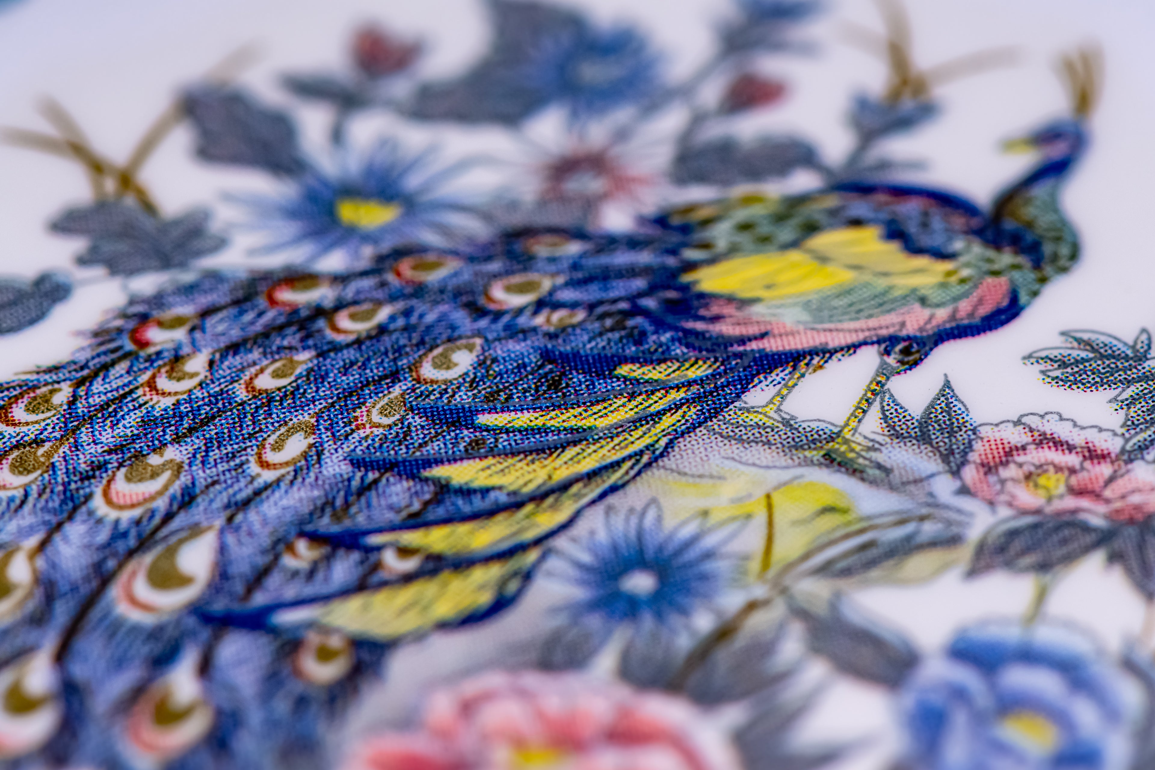 A close-up of a colorful embroidered peacock with blue, yellow, green, and pink details, surrounded by floral motifs on a white background.