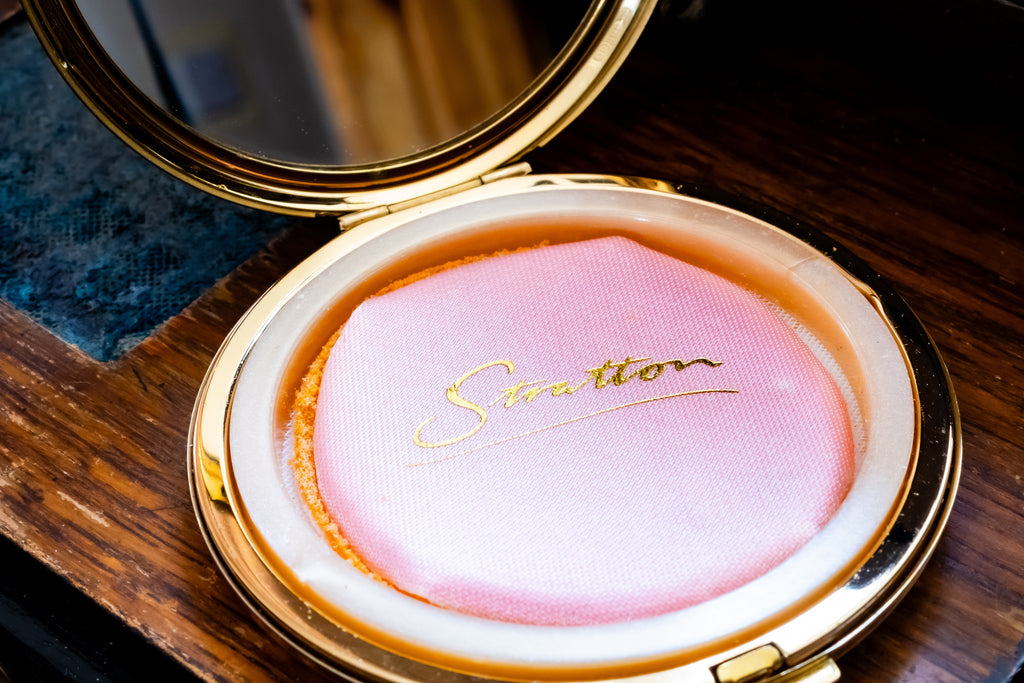 An open round gold compact containing a pink pressed powder with the brand name 'Stratton' in gold script, resting on a dark wooden surface.