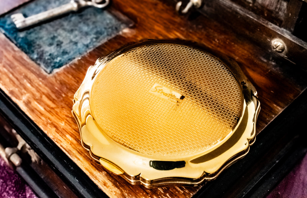 A close-up of a round gold-toned Stratton powder compact with a textured center and scalloped edge, resting on a dark wooden surface with a vintage key nearby.