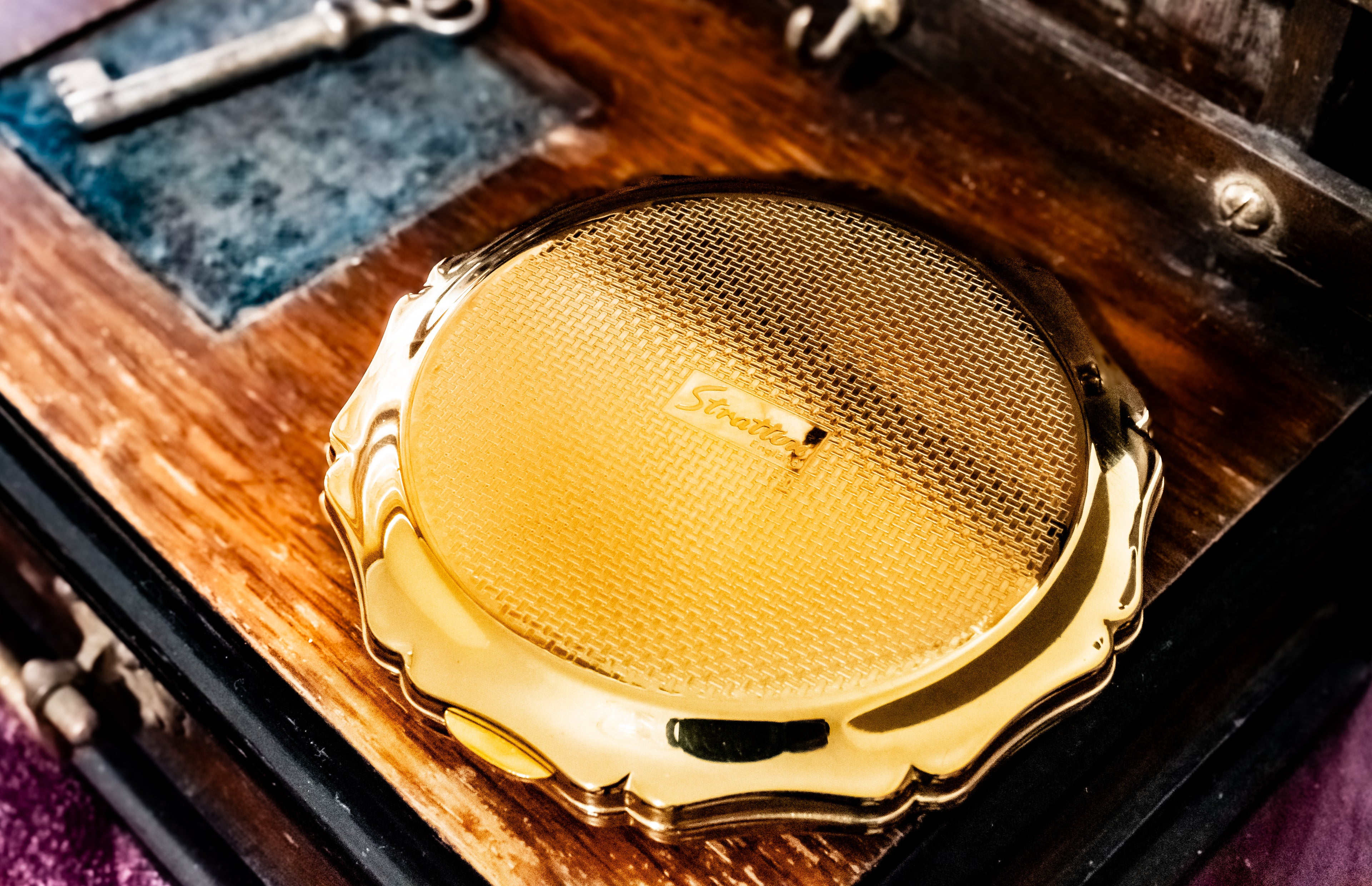 A close-up of a round gold-toned Stratton powder compact with a textured center and scalloped edge, resting on a dark wooden surface with a vintage key nearby.