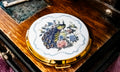 A round compact mirror with a gold-tone rim and a white center decorated with a detailed peacock and floral illustration, resting on a polished wooden surface.