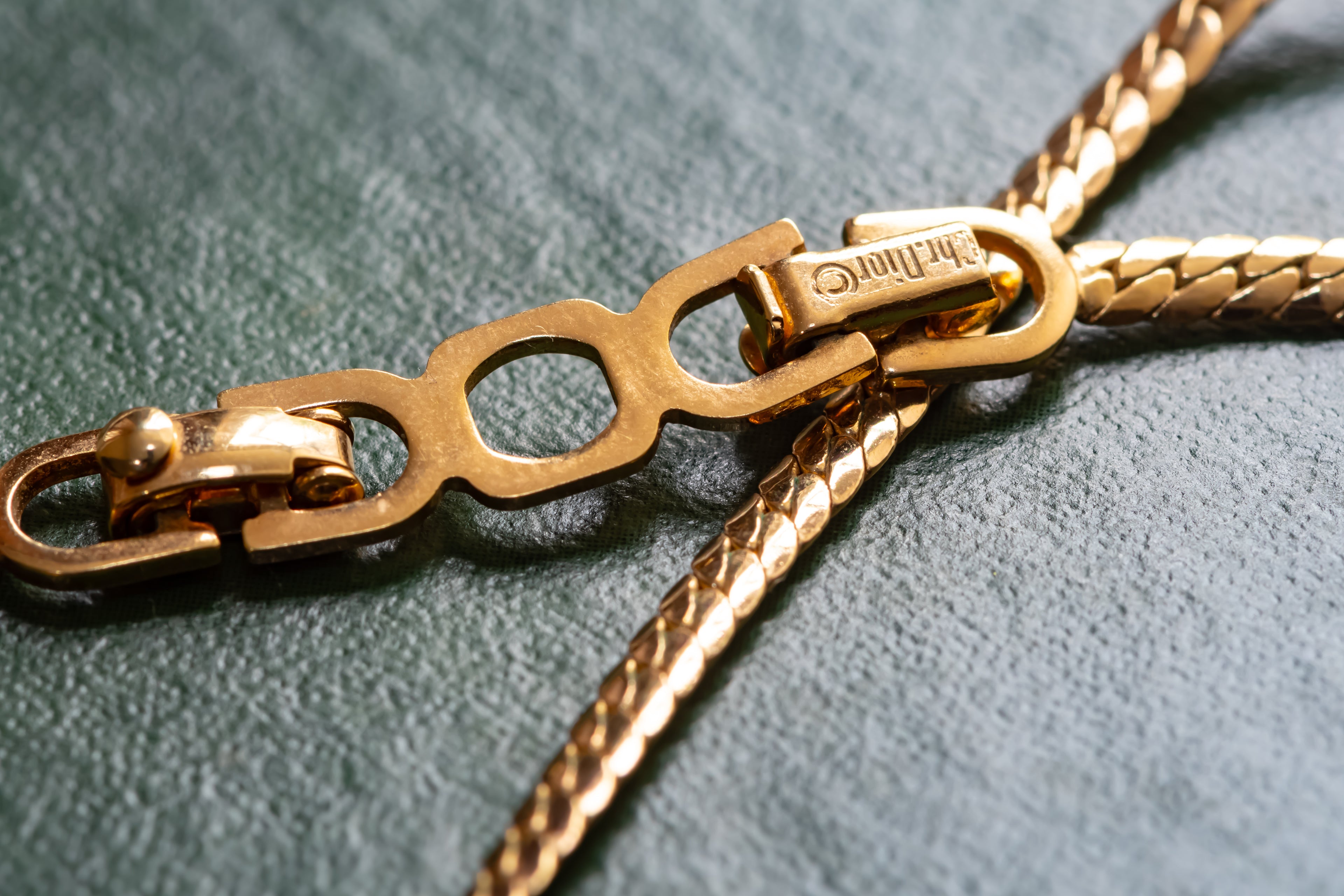 Close-up of a gold-tone chain necklace with a distinctive three-ring clasp and a braided section, resting on a textured dark green surface. The brand name 'Christian Dior' is engraved on the clasp.