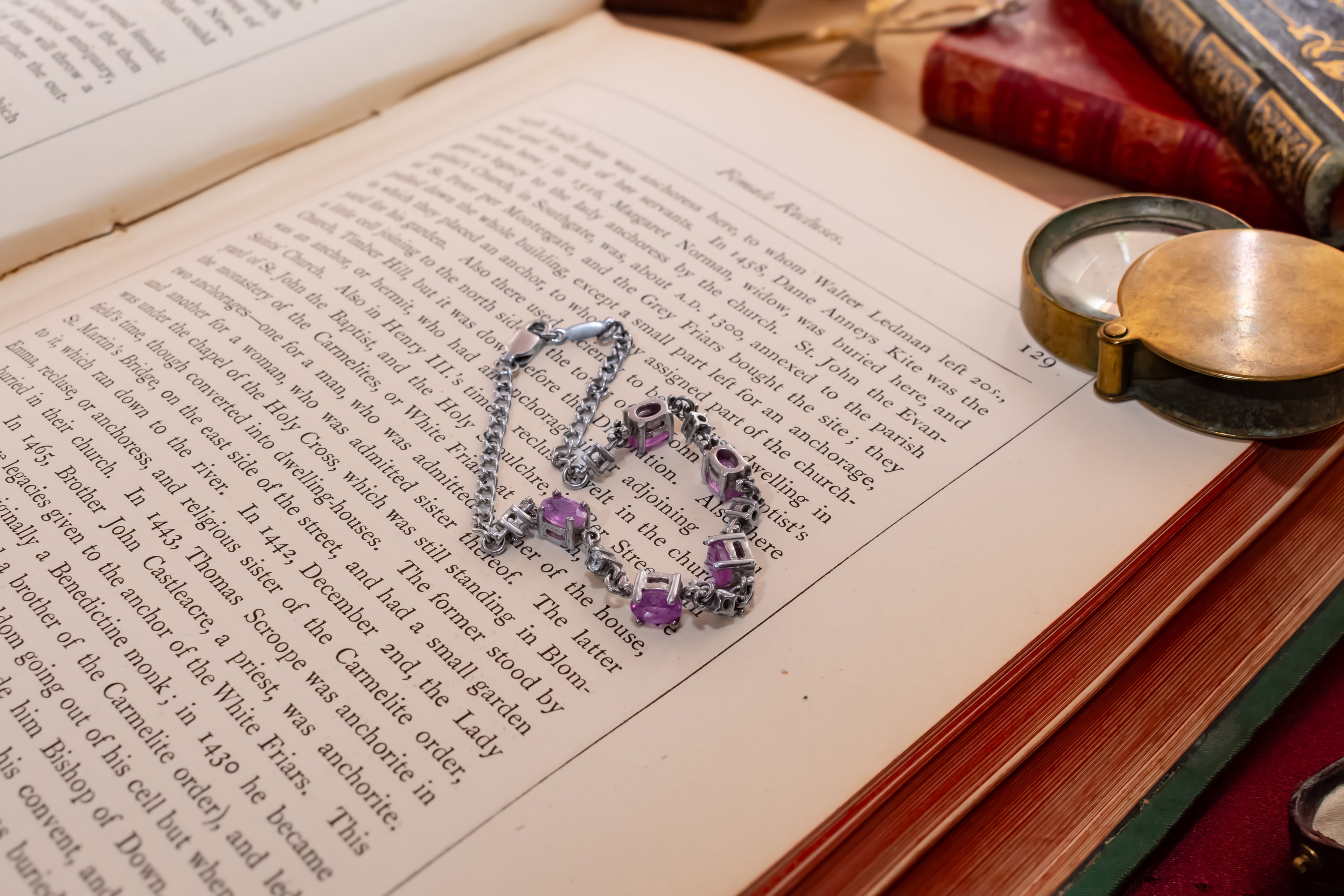 A silver-toned heart-shaped necklace with purple gemstone accents lies on the pages of an open book, surrounded by vintage books and a brass pocket watch.