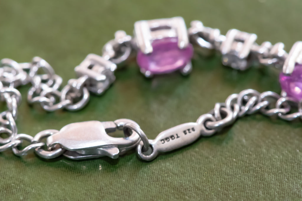 Silver chain bracelet with oval purple gemstones and a lobster clasp, shown close-up on an olive green textured background. The clasp is engraved with '925 TITAN'.
