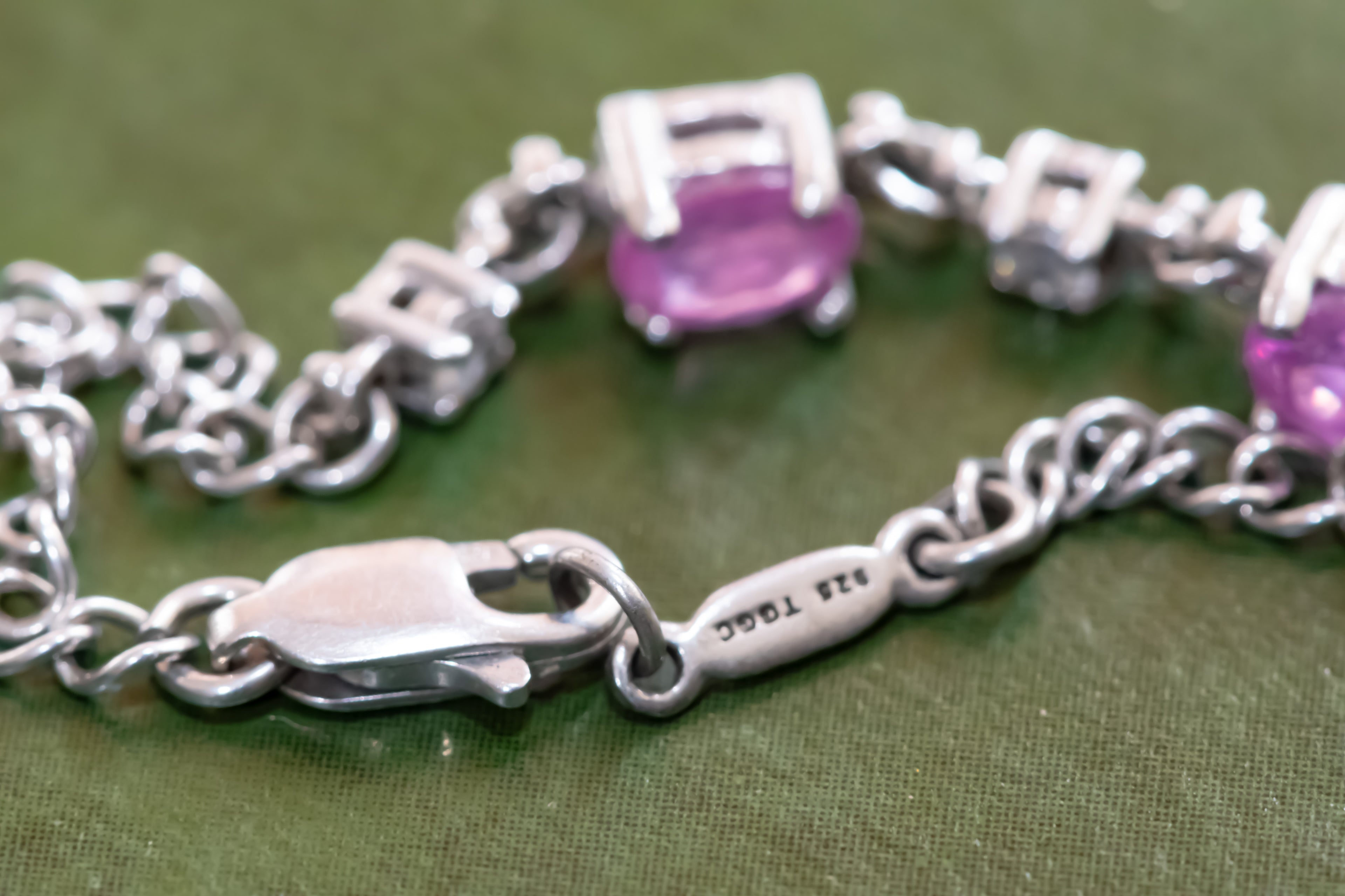 Silver chain bracelet with oval purple gemstones and a lobster clasp, shown close-up on an olive green textured background. The clasp is engraved with '925 TITAN'.