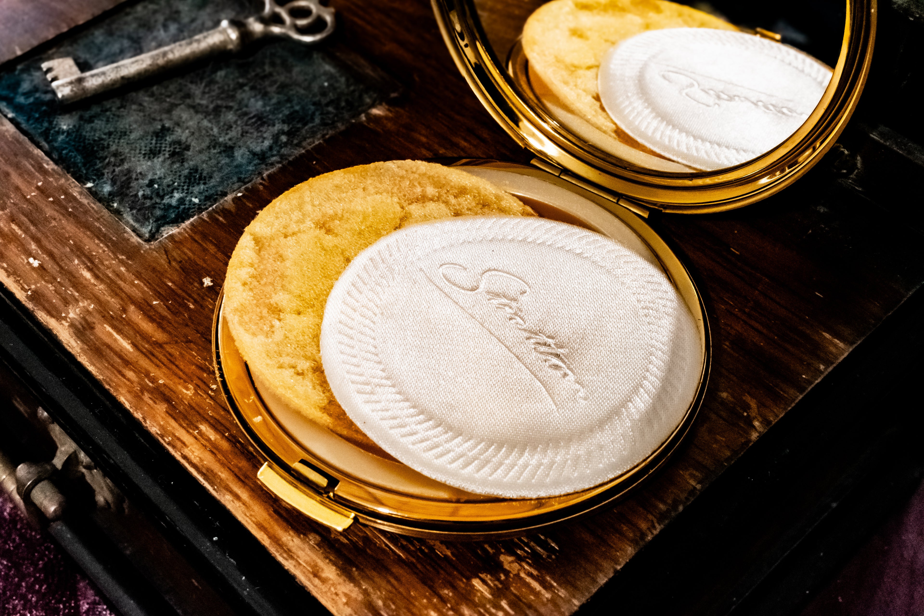 A gold compact mirror holding a round white cotton pad with an embossed logo, resting on a vintage wooden box next to an antique key.