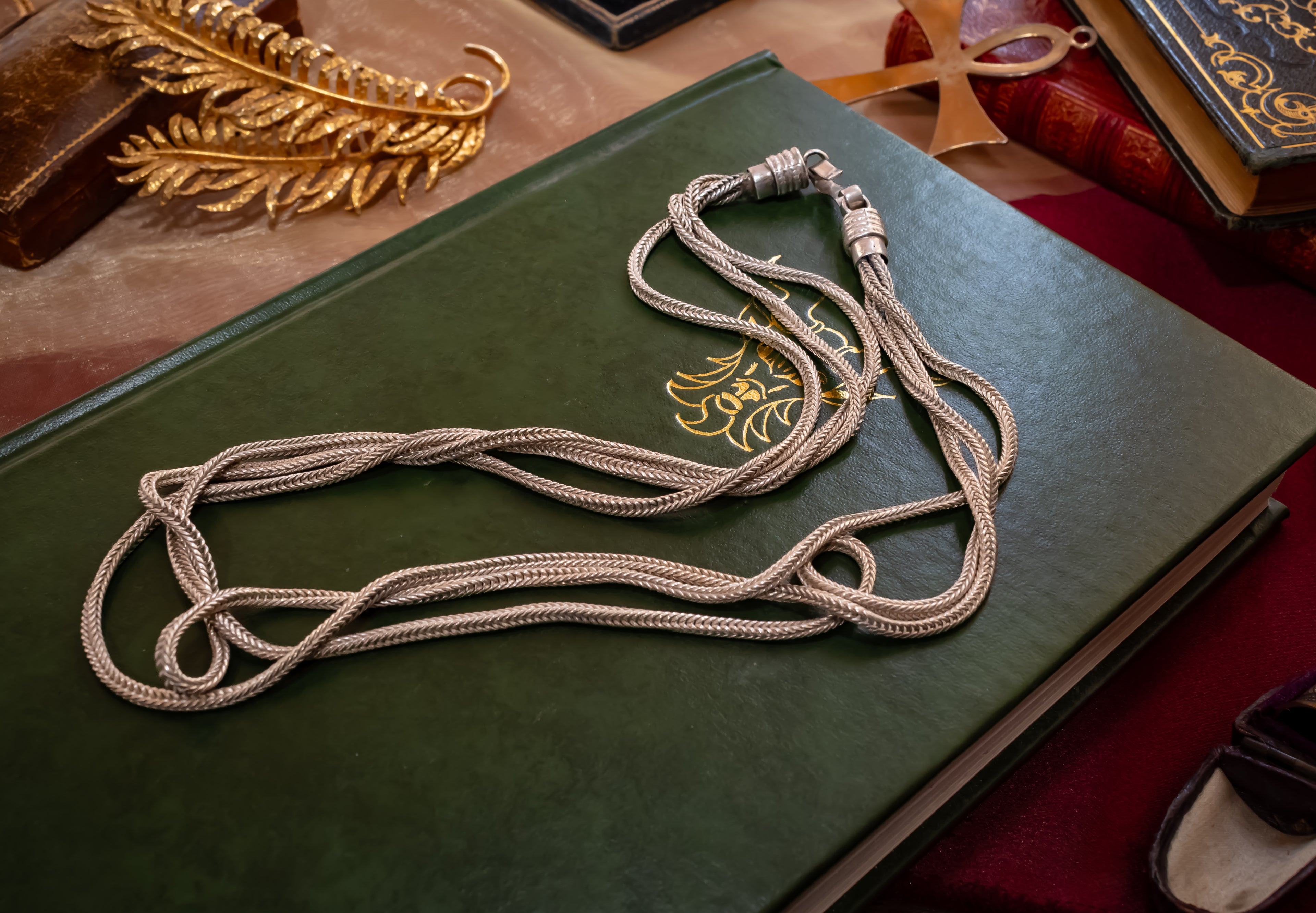 A silver braided cord necklace with a silver toggle clasp, draped over a dark green leather-bound book with gold lettering.