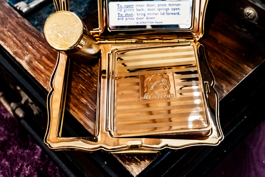 A gold-toned vintage-style compact case opened on a dark wooden surface, showing a striped mirror interior with an instruction label and a round gold seal attached to the side.