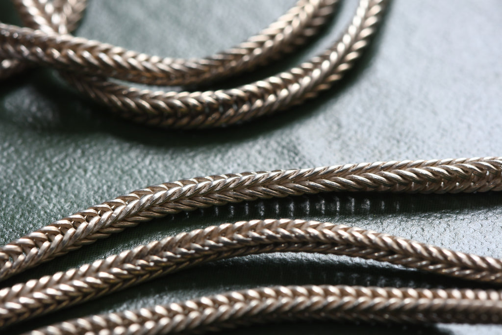 Close-up of braided chain necklace  in a silver-toned finish, draped across a dark textured surface.