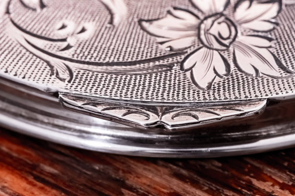 Close-up of a silver-toned metal container lid with engraved floral detailing and a textured pattern, resting on a dark wooden surface.