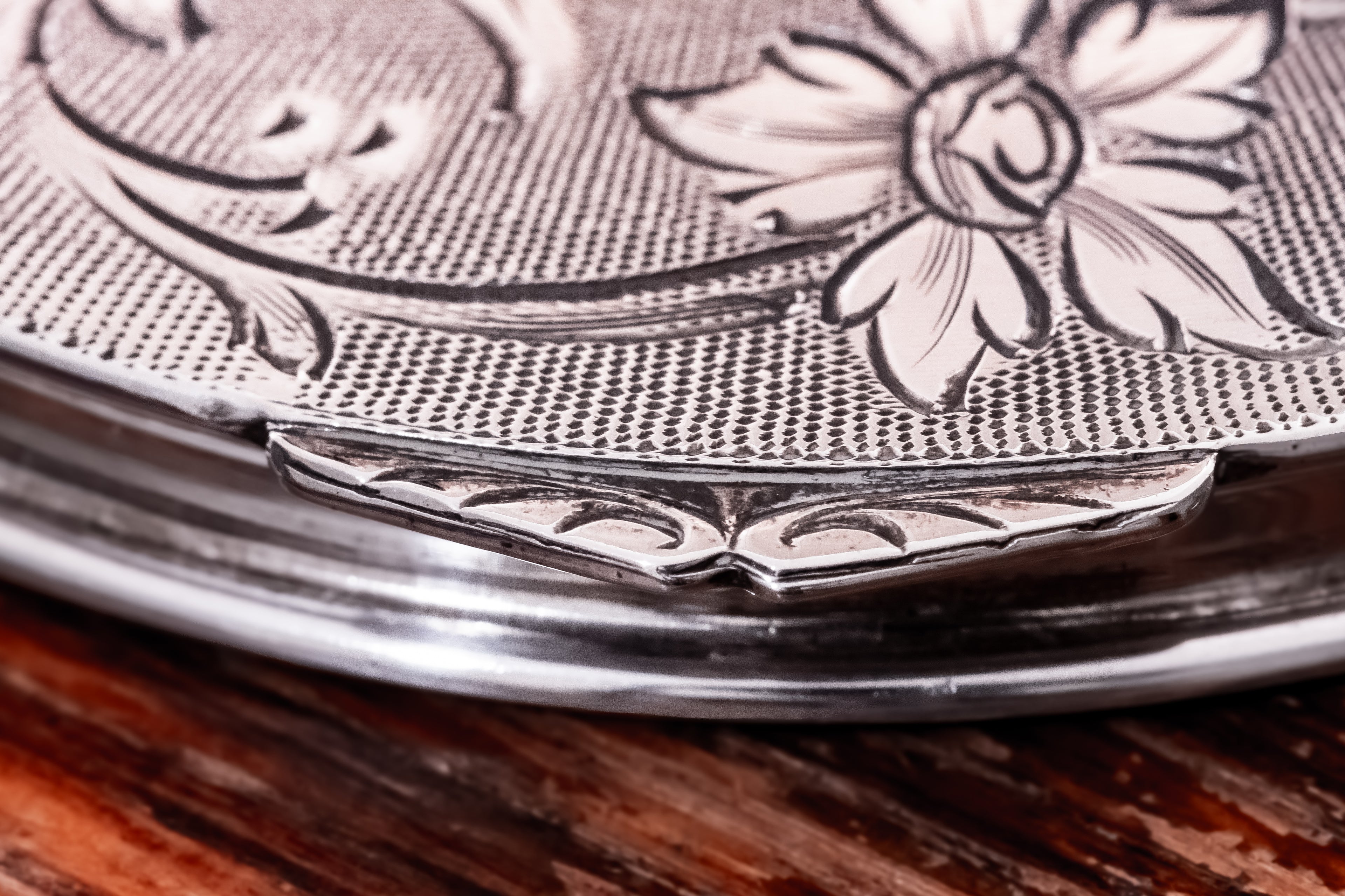 Close-up of a silver-toned metal container lid with engraved floral detailing and a textured pattern, resting on a dark wooden surface.
