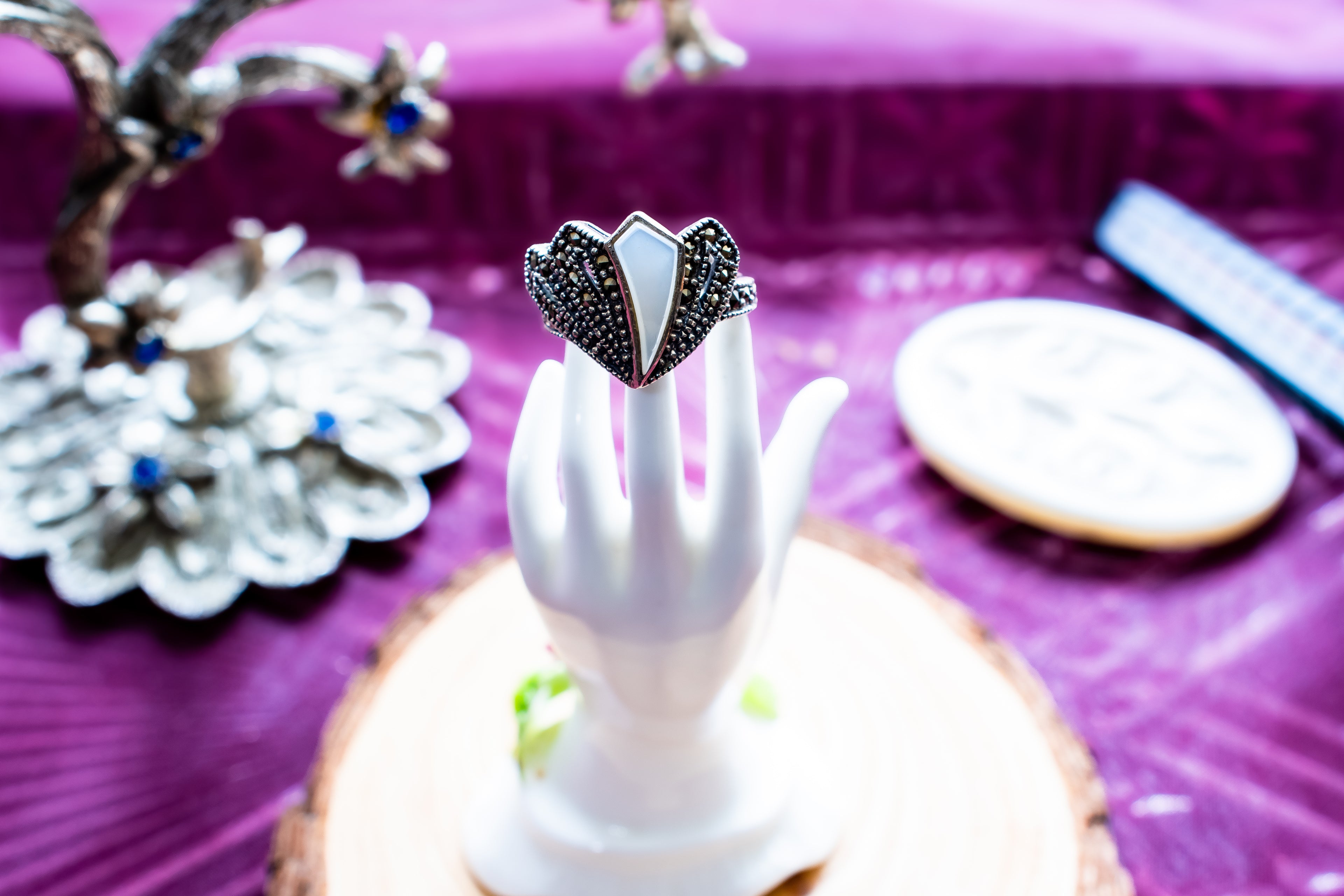 A heart-shaped silver ring with black pavé stones and a white inlay, displayed on a white hand-shaped stand on a wooden slice over a purple surface.