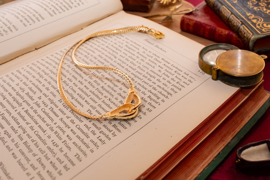 A gold chain necklace with a heart-shaped pendant is displayed on an open book, surrounded by vintage books and a small brass locket.