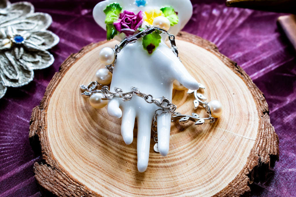 A silver-toned chain necklace with white pearl-like beads draped over a white ceramic hand-shaped display, set on a round wood slice over a purple fabric background.