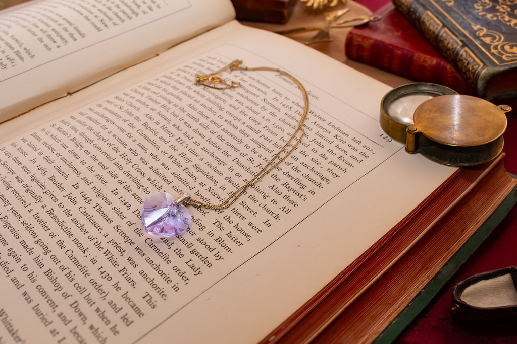 A gold chain necklace with a faceted light purple crystal pendant rests on the pages of an open book, surrounded by vintage books and a brass pocket mirror.