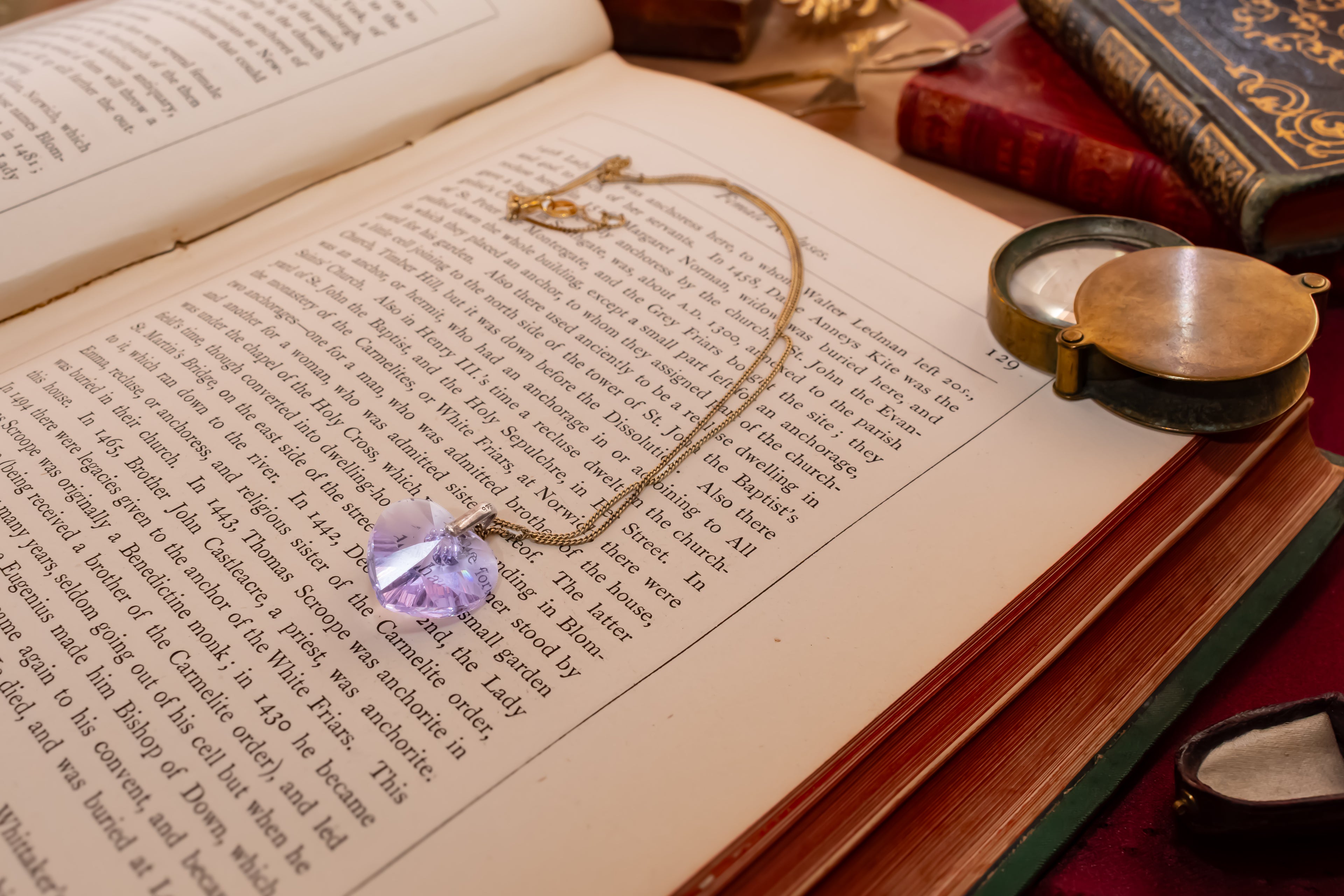 A gold chain necklace with a faceted light purple crystal pendant rests on the pages of an open book, surrounded by vintage books and a brass pocket mirror.