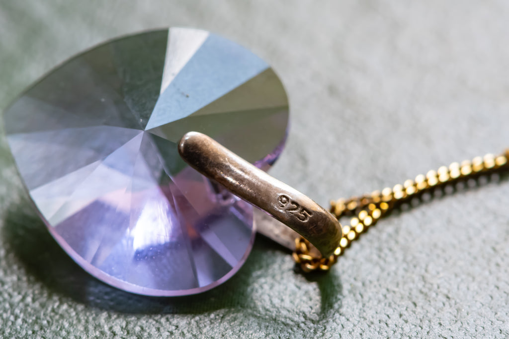 A close-up of a faceted heart-shaped clear crystal pendant with a gold chain and a gold ring clasp engraved with '925'.
