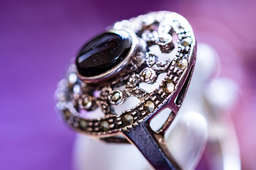An ornate silver-toned ring with a large oval black stone and small clear stones in a filigree setting, photographed close-up against a purple background.