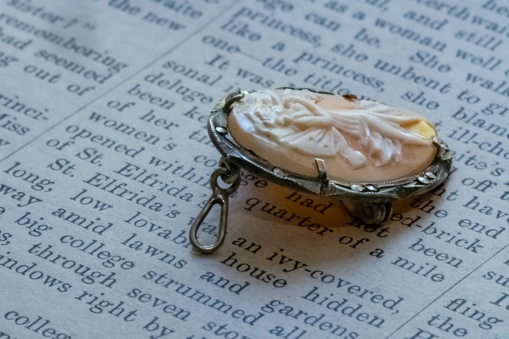 A vintage-style carved shell pendant with a silver-toned metal frame and loop, resting on an open book page with printed text.