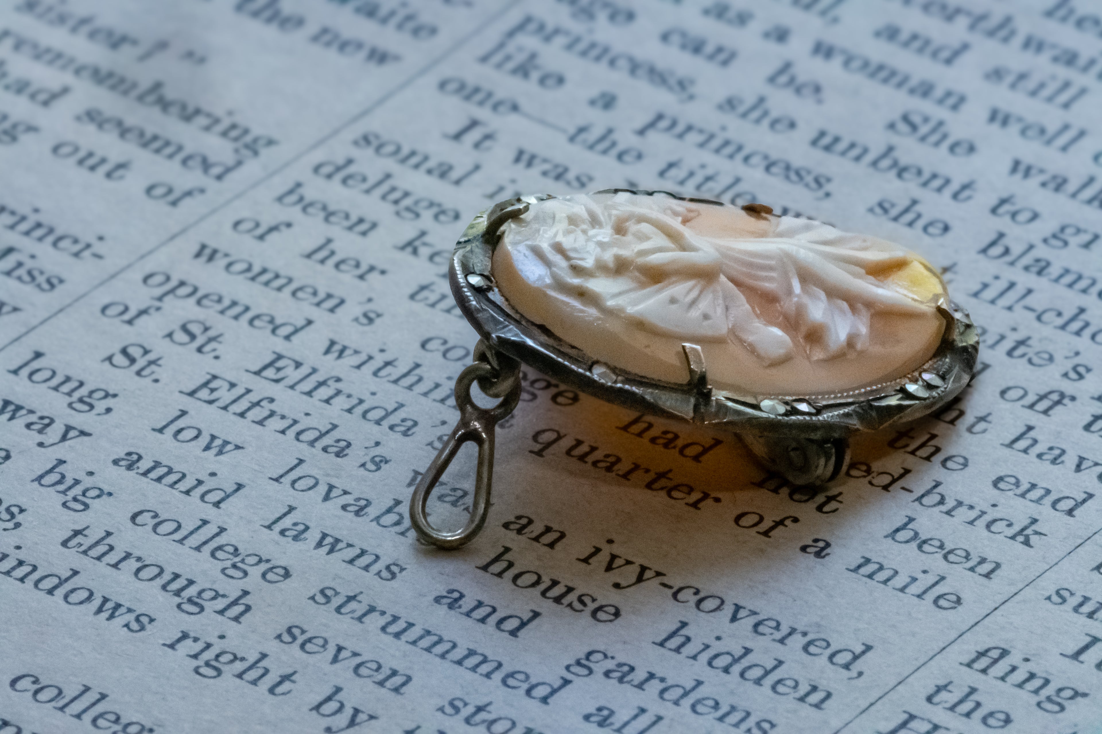 A vintage-style carved shell pendant with a silver-toned metal frame and loop, resting on an open book page with printed text.