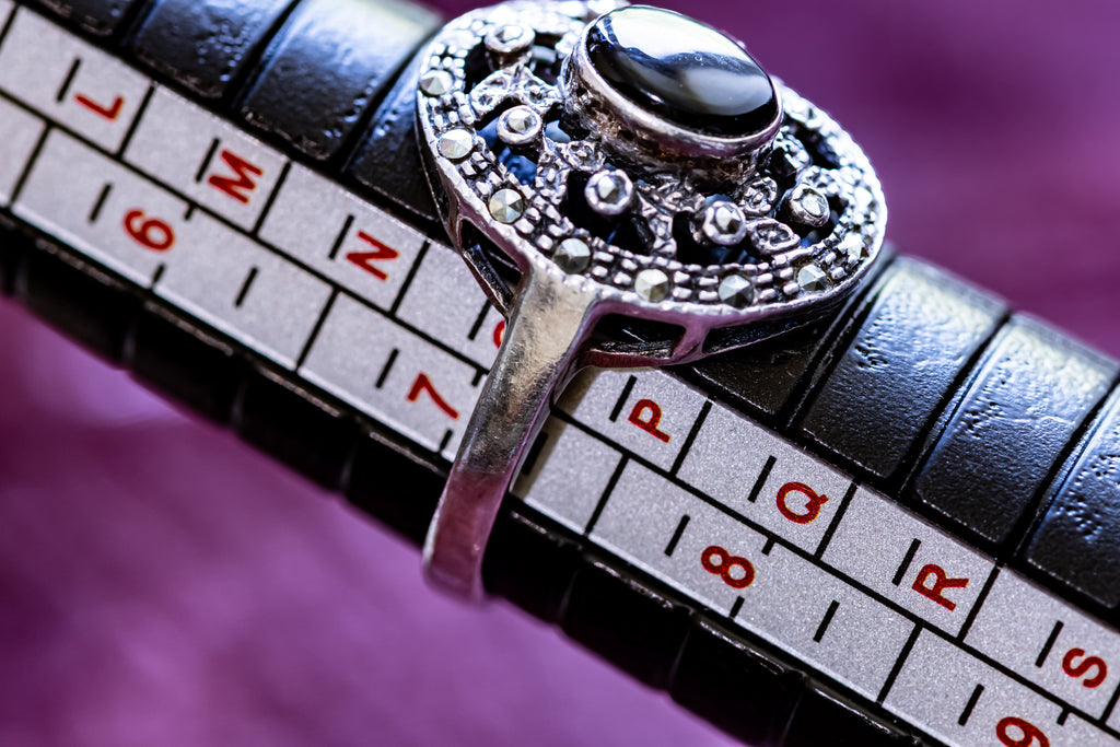 A silver-toned ring with a black oval stone and ornate filigree detailing, shown on a black leather sizing ring with a white size scale.