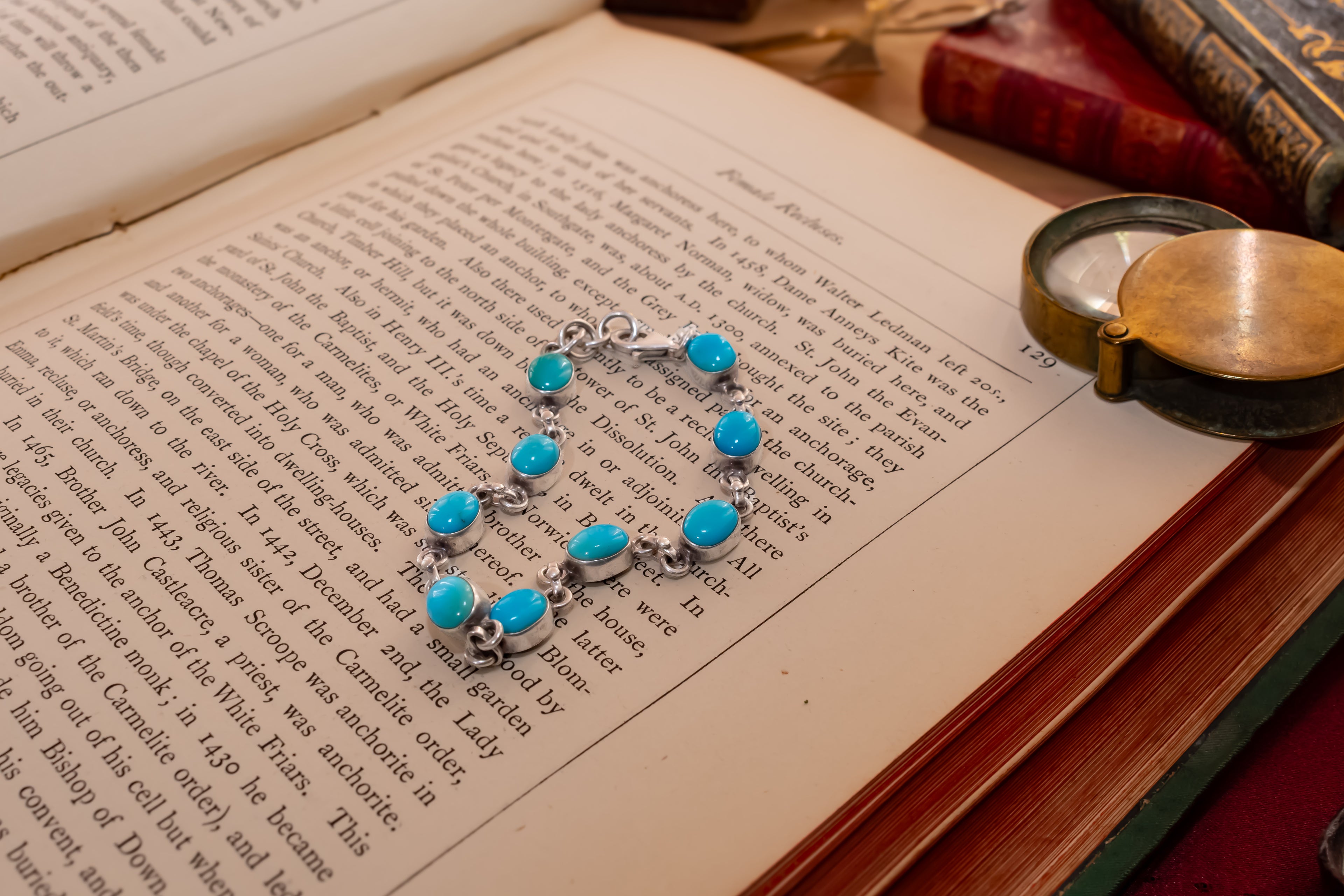 A silver-toned bracelet with nine round turquoise-blue stones is laid across the pages of an open book, with vintage books and a brass pocket mirror in the background.
