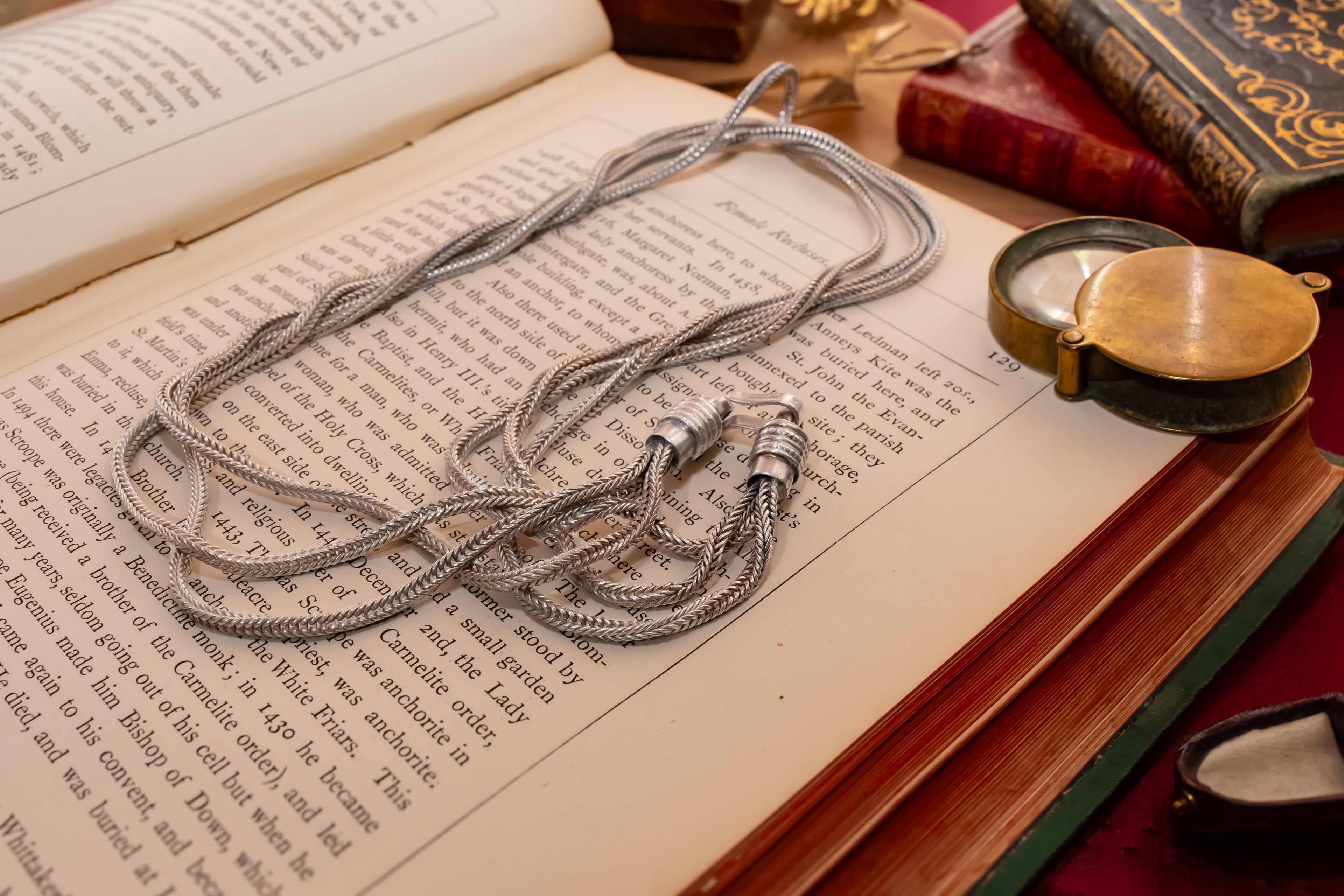 A silver braided chain necklace with a hook like clasp, laid across the pages of an open vintage book. The scene includes old leather-bound books and a brass pocket watch nearby, creating a warm, antique aesthetic.