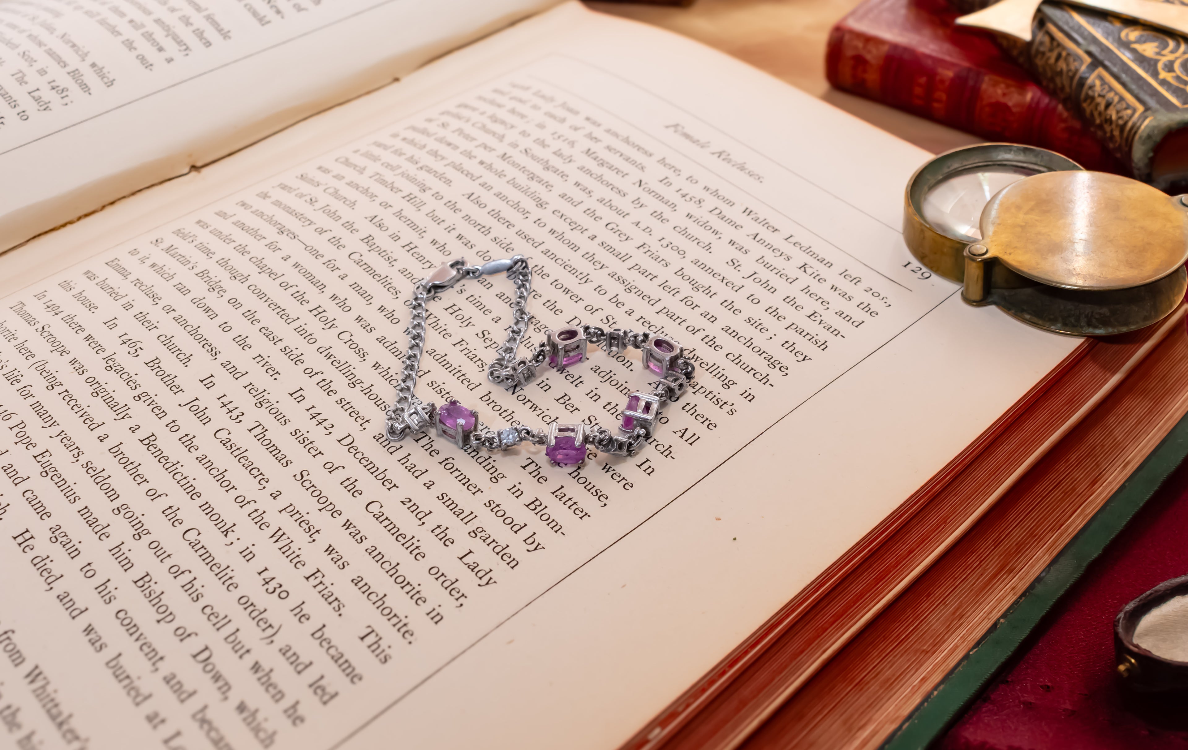 A silver-toned link bracelet with purple gemstone accents lies on the pages of an open book, surrounded by vintage books and a brass pocket mirror.