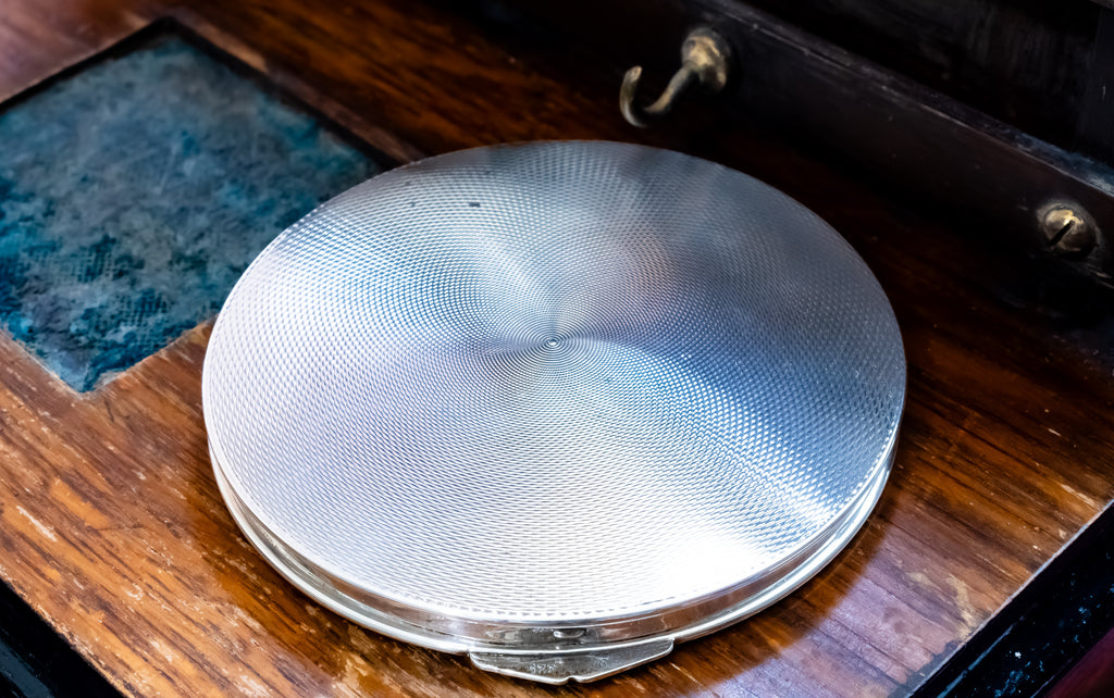 A round silver metal compact case with a textured, brushed finish, resting on a dark brown wooden surface.