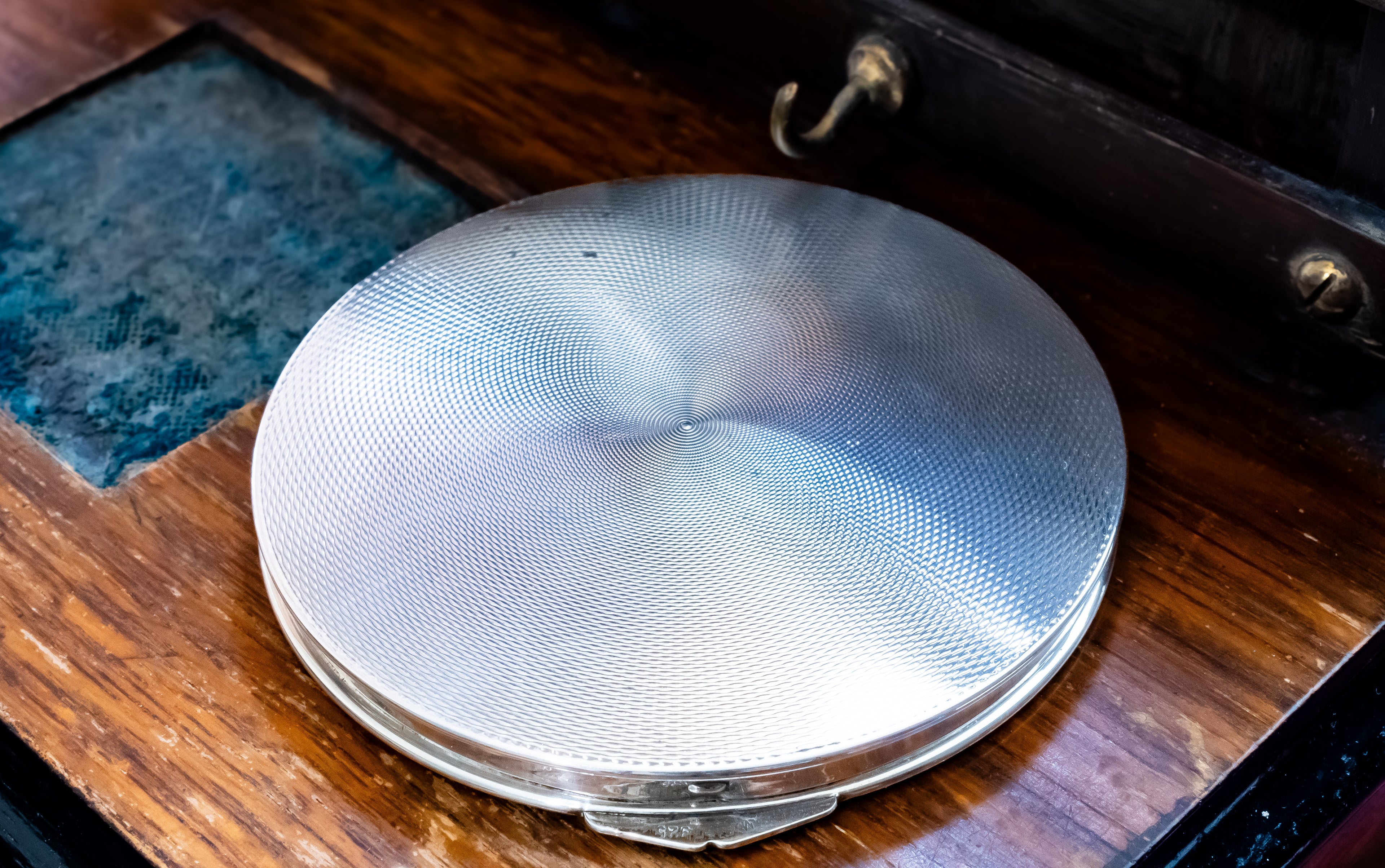 A round silver metal compact case with a textured, brushed finish, resting on a dark brown wooden surface.