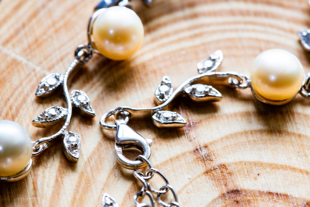 Close-up of a silver-toned leafy vine jewelry piece with round white pearls and small clear stones, resting on a wood surface.