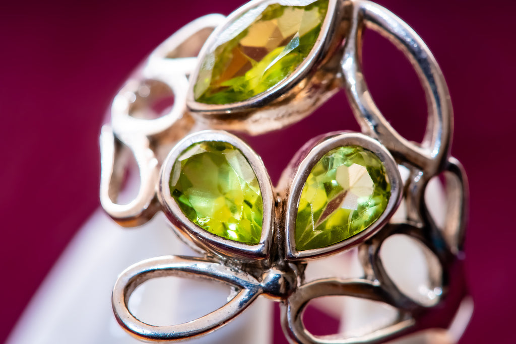 A close-up of a silver-toned ring featuring three faceted green gemstones in a leaf-like openwork design, set against a deep magenta background.