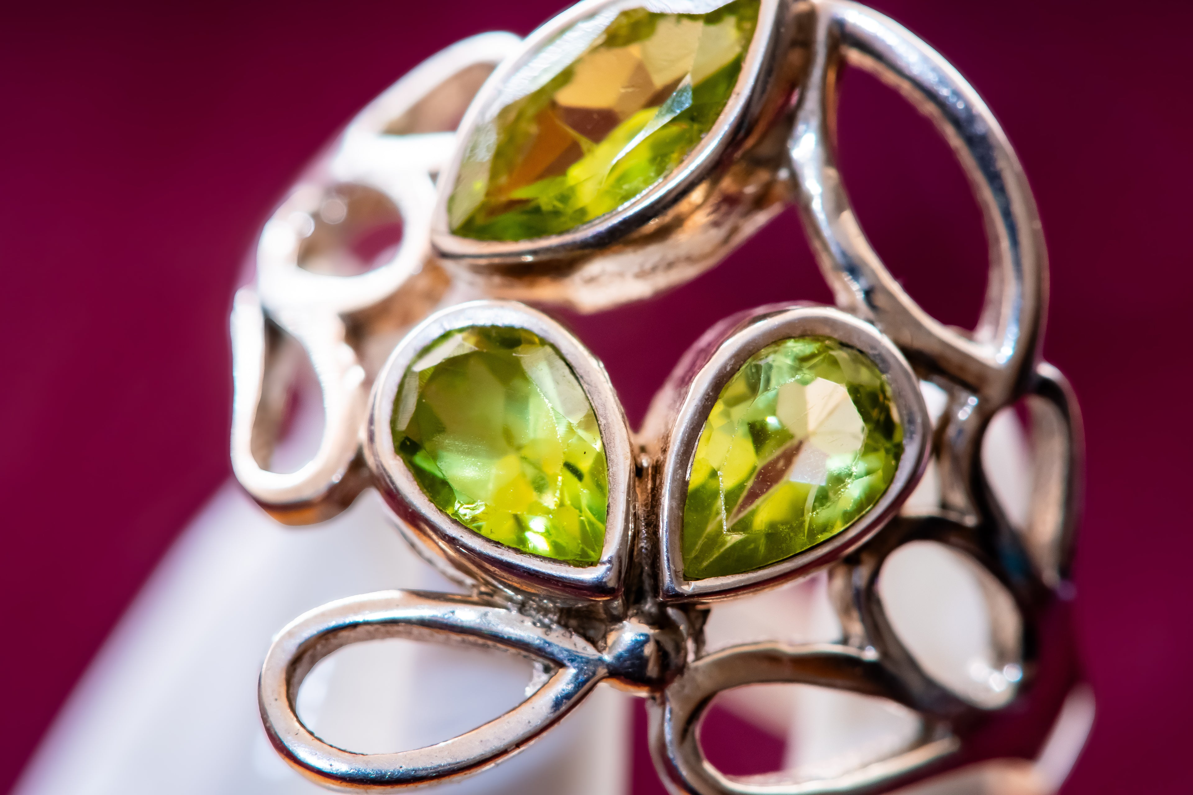 A close-up of a silver-toned ring featuring three faceted green gemstones in a leaf-like openwork design, set against a deep magenta background.