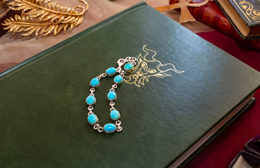 A silver chain bracelet with oval turquoise-blue stones, displayed on a dark green book with gold ornamental detailing.