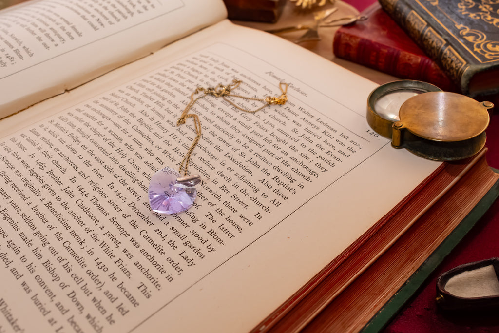 A heart-shaped light purple crystal pendant on a gold chain, resting on the pages of an open book surrounded by vintage books and a brass pocket mirror.