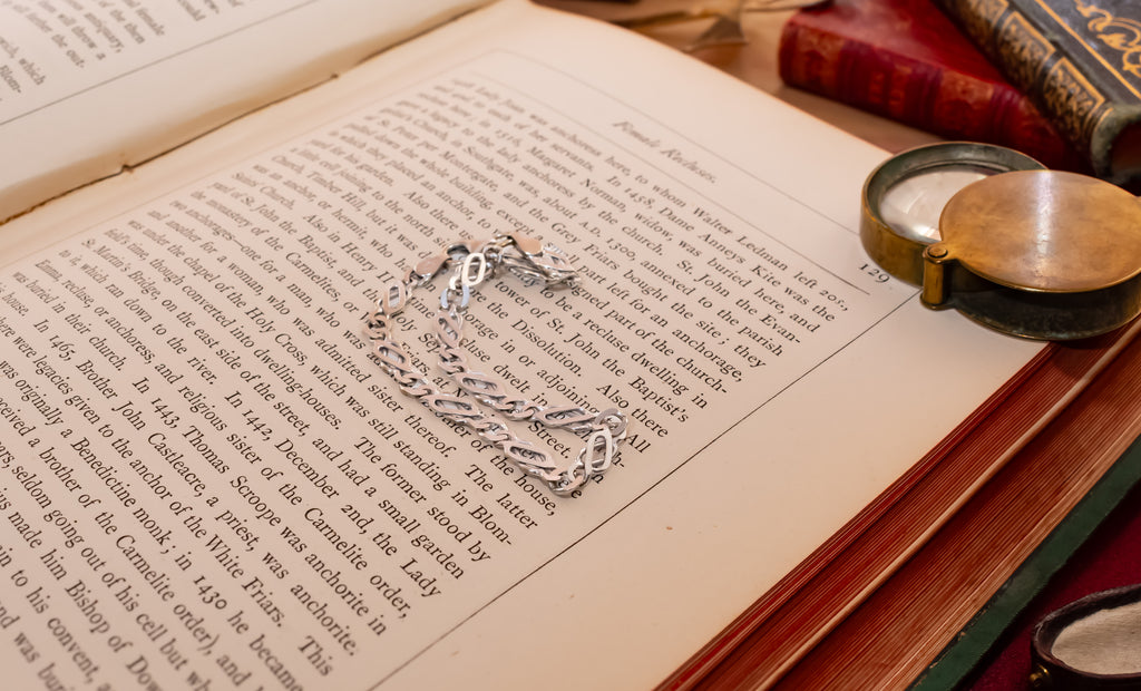 A silver-toned chain bracelet with a small pendant rests on the open pages of an antique book, surrounded by old books and a brass pocket mirror on a wooden surface.