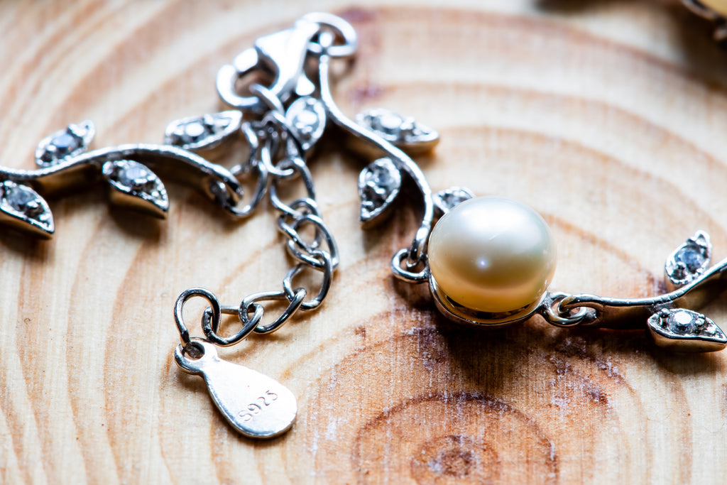Silver branch-shaped charm with leaf details, clear rhinestones, and a round cream pearl, displayed on a wooden surface.