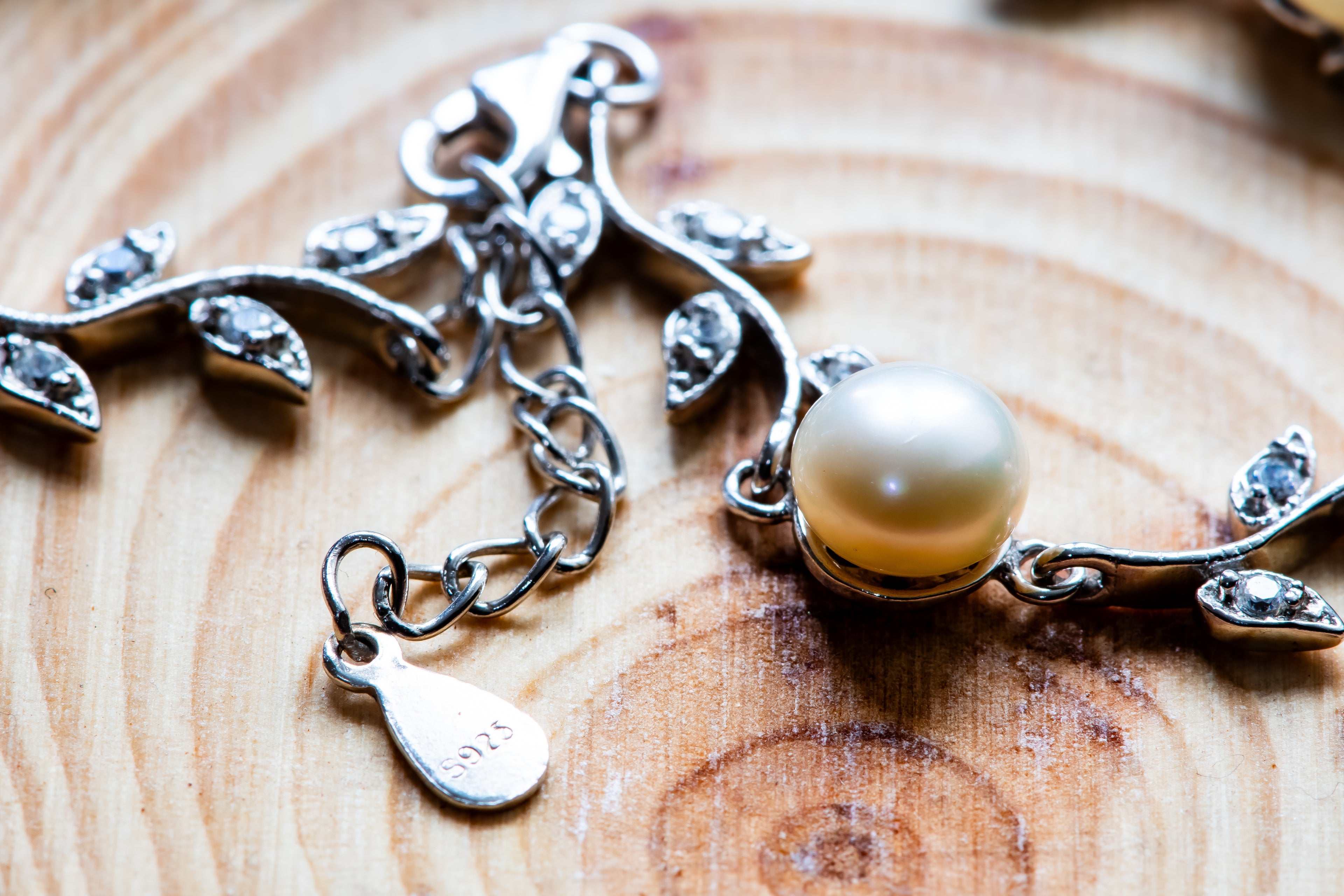 Silver branch-shaped charm with leaf details, clear rhinestones, and a round cream pearl, displayed on a wooden surface.
