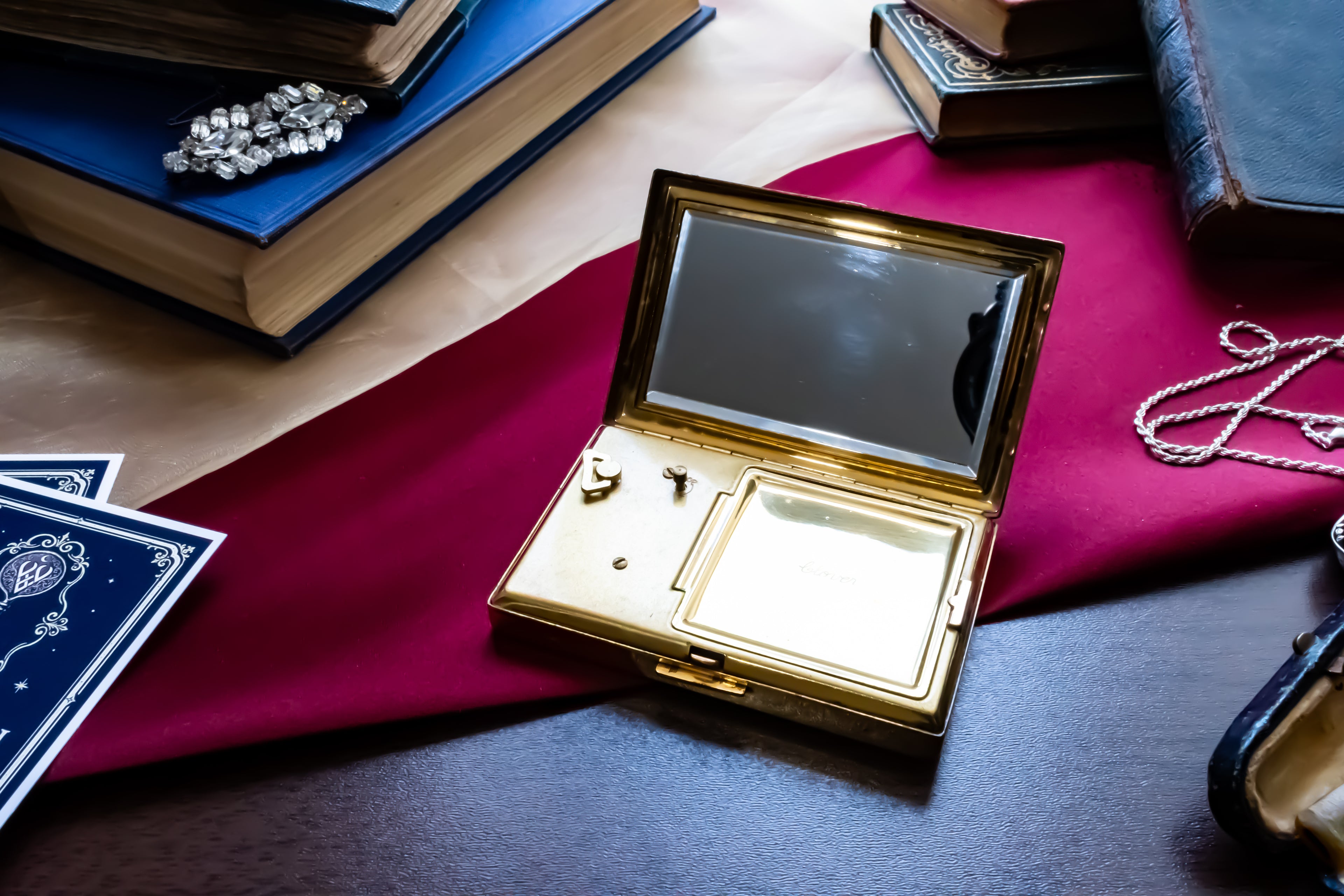 An open gold-toned musical powder compact with a mirror and turn key pieces inside, styled on a dark tabletop with a red cloth, books, cards, and a necklace nearby.