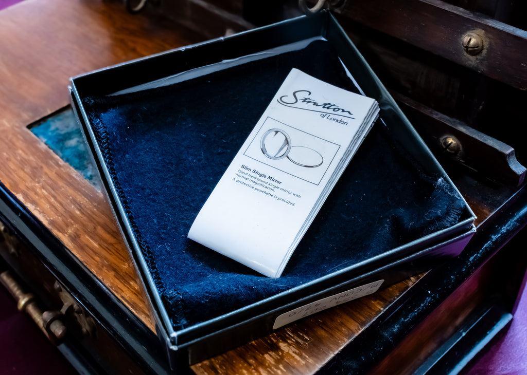 A white paper card with the 'Sutton of London' brand logo and an illustration of a single round gemstone, placed on a dark blue velvet-lined black jewelry box inside an antique wooden chest.
