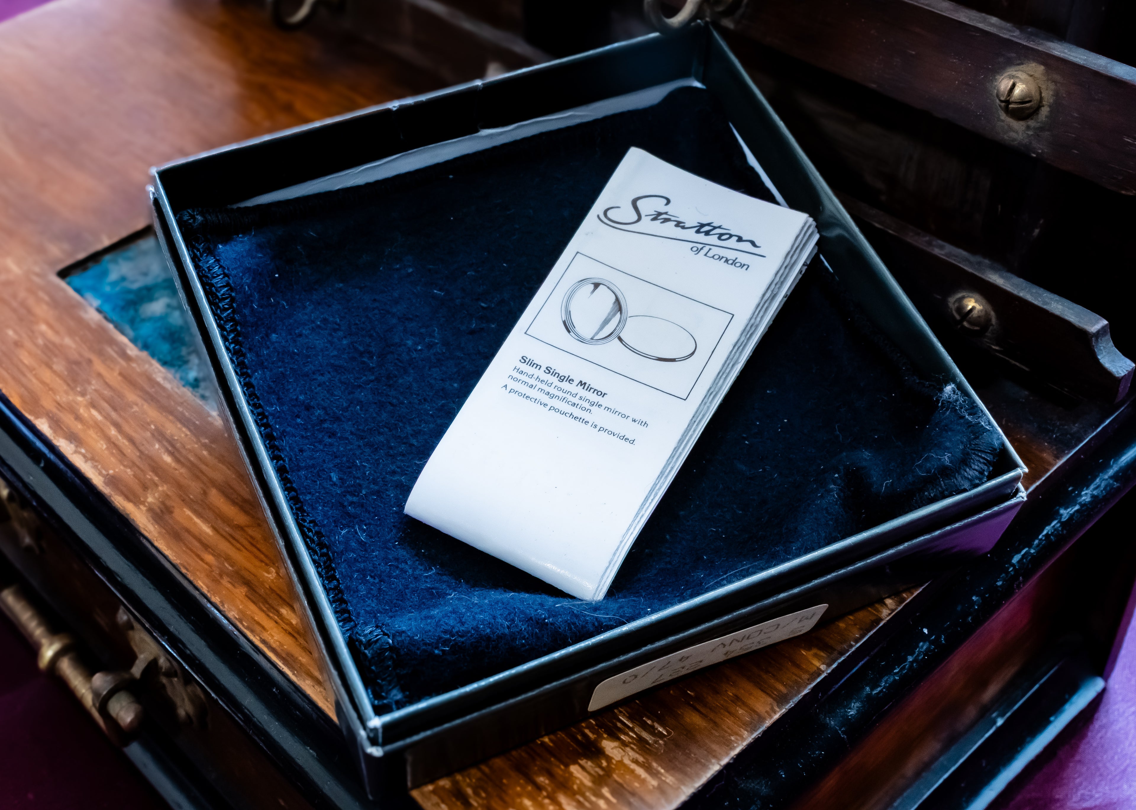 A white paper card with the 'Sutton of London' brand logo and an illustration of a single round gemstone, placed on a dark blue velvet-lined black jewelry box inside an antique wooden chest.